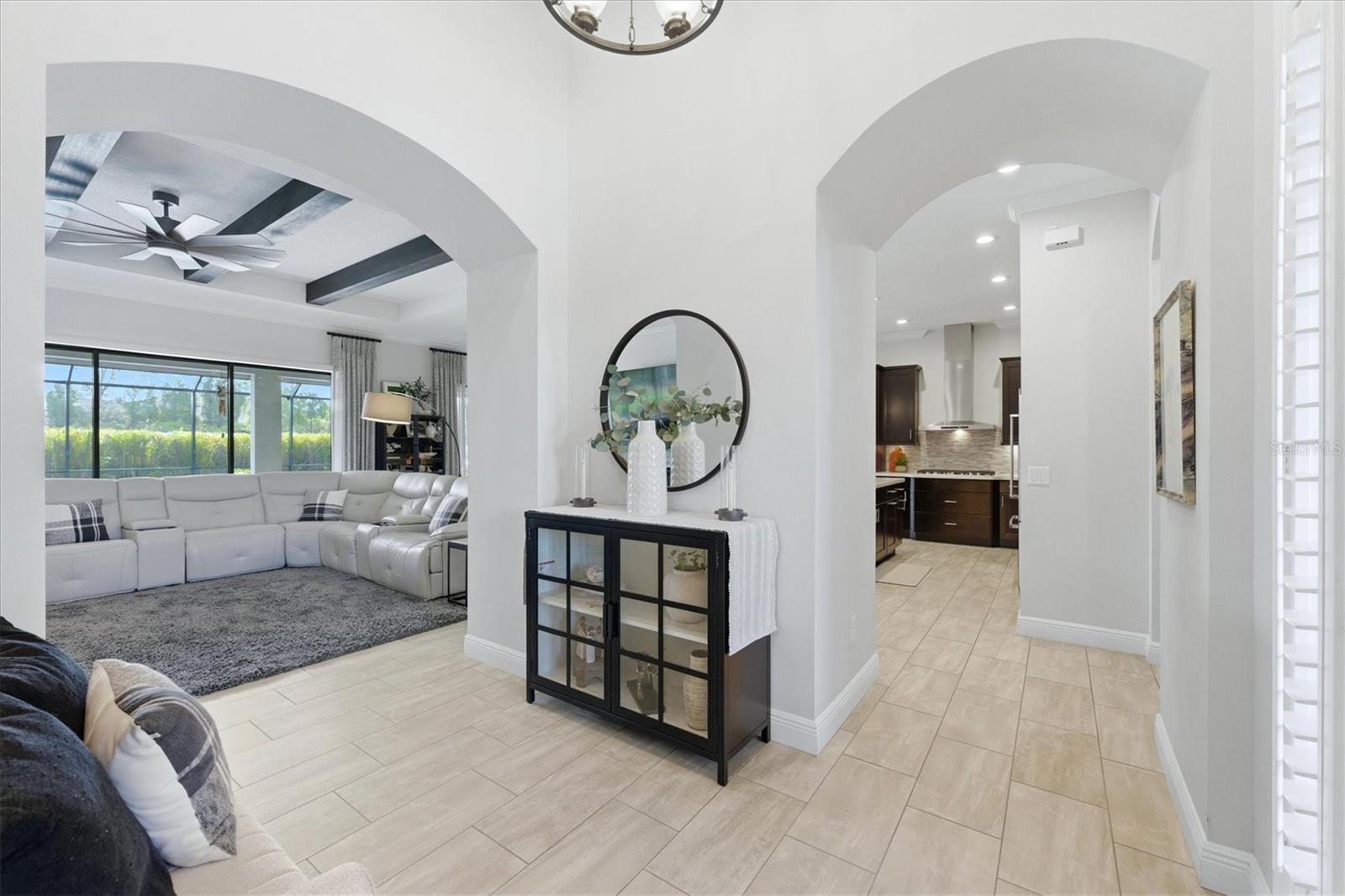 Foyer with arched openings to a living room and kitchen, pale tile floors, and a black console beneath a round mirror.
