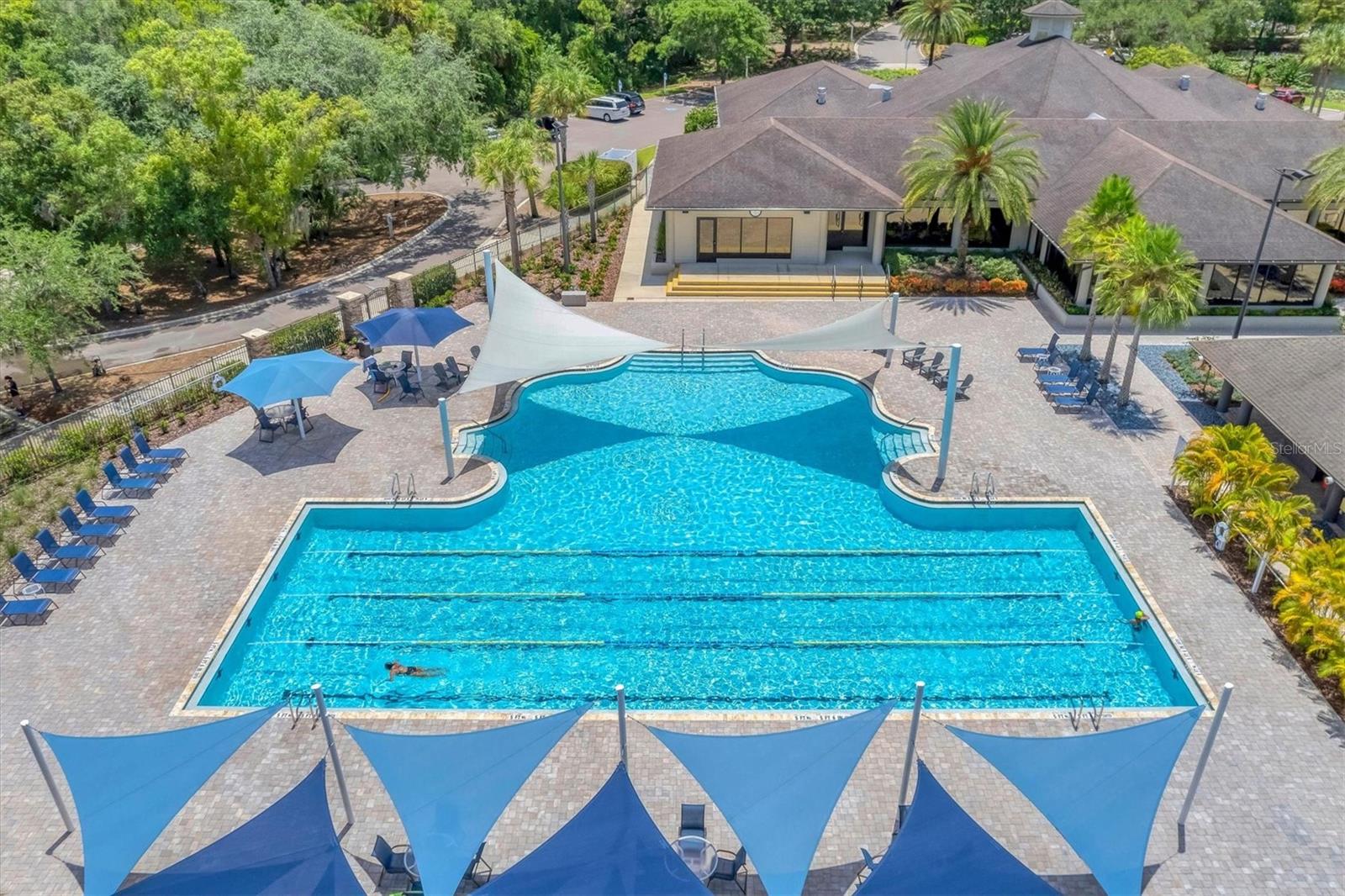 Outdoor swimming pool with blue shade sails and a clubhouse surrounded by palm trees.