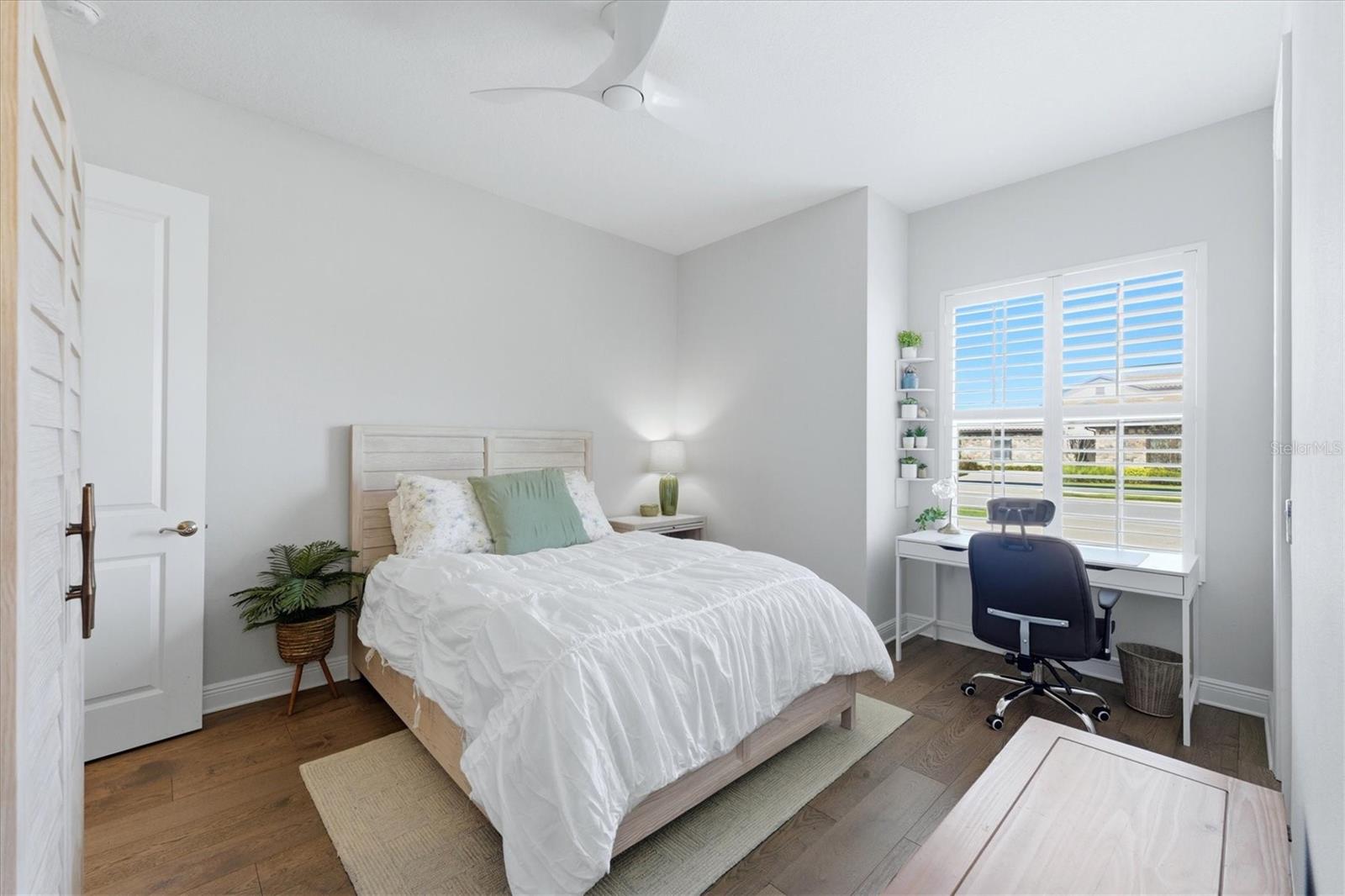 Staged bedroom with white bedding, a desk by shuttered windows, and warm wood floors.