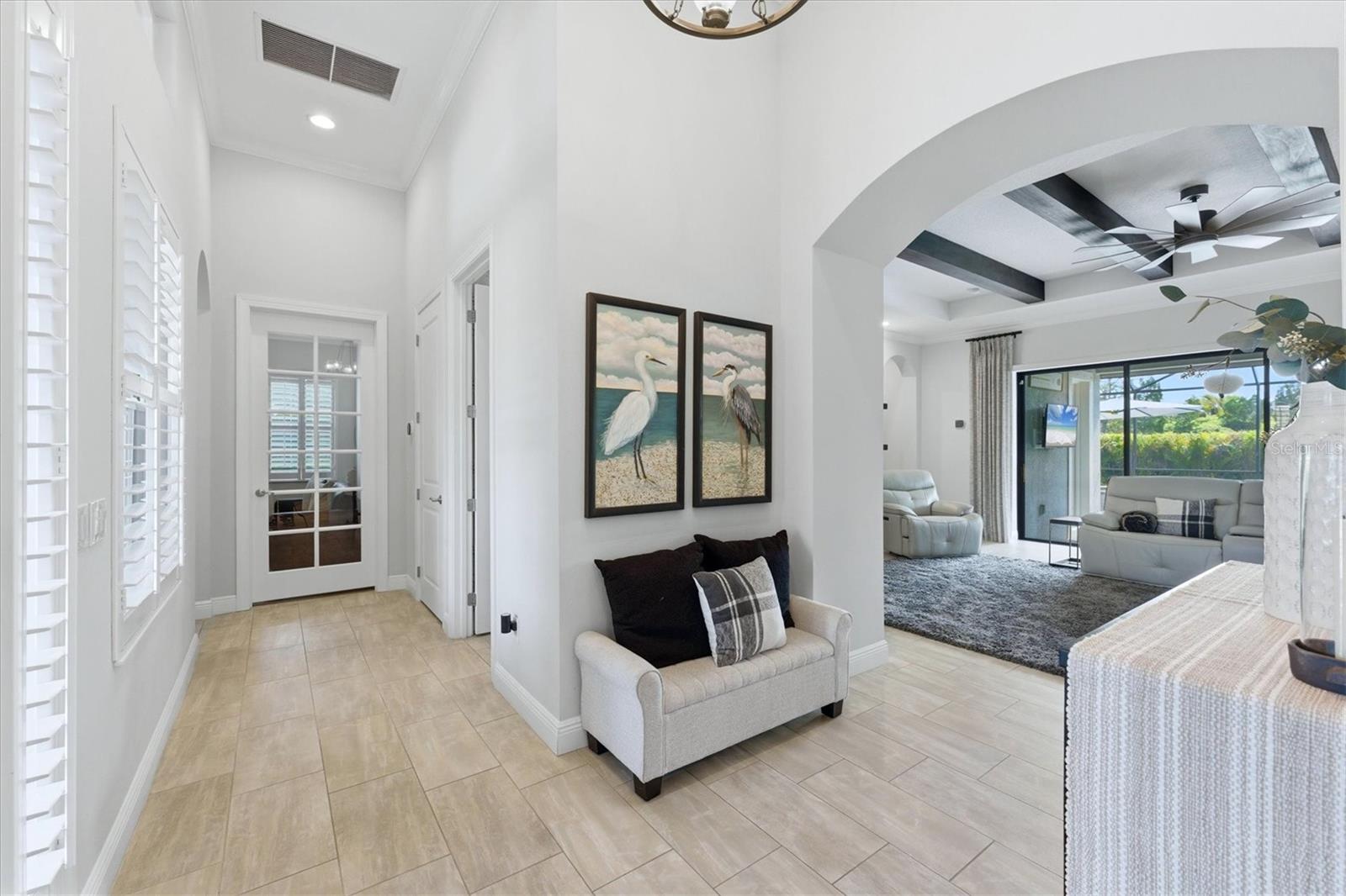Foyer opening to a furnished living room with tile floors, plantation shutters, and sliding glass doors.