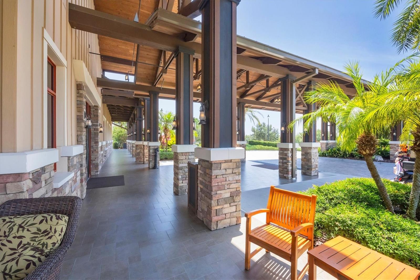 Covered patio with timber beams, stone columns, tiled flooring, and seating beside palm trees.