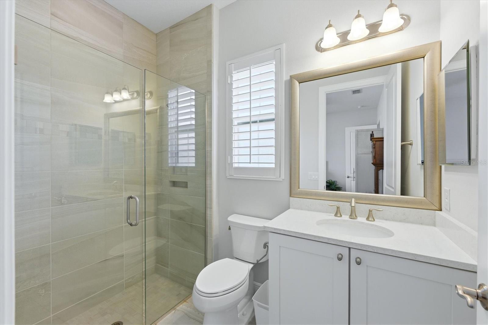 Bathroom with a tiled glass shower, white vanity, gold-toned fixtures, and shuttered window.