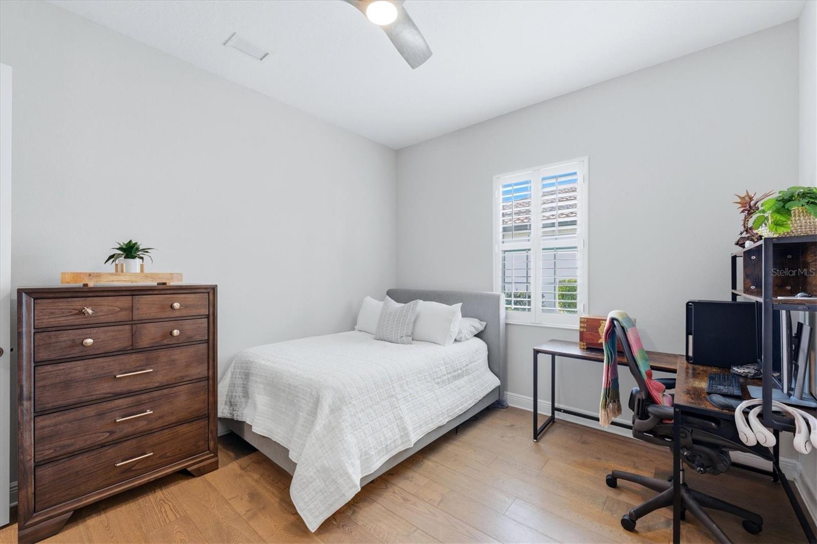 Bedroom with a white-covered bed, wood dresser, and desk beneath plantation-shuttered window.
