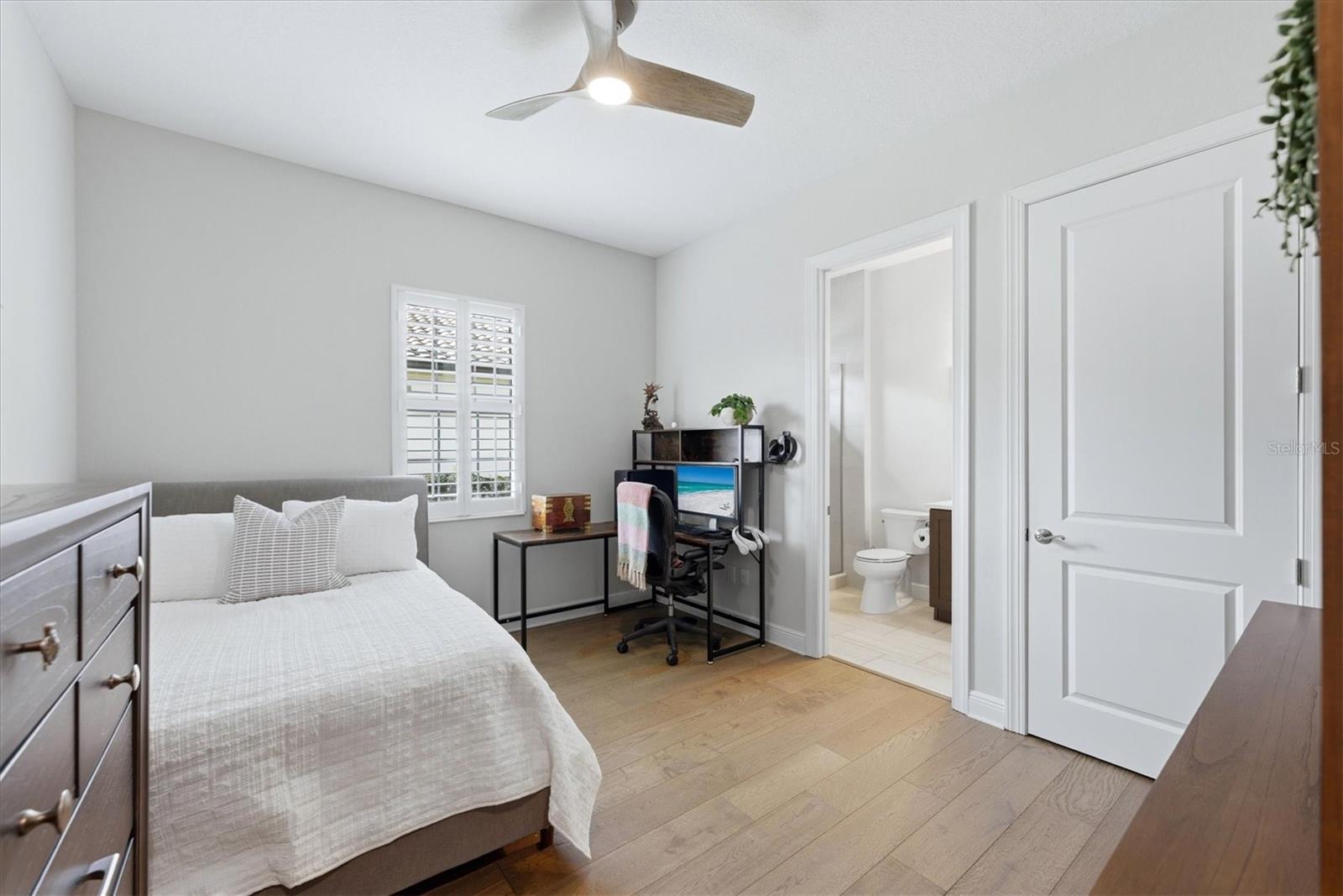 Bedroom with a white bed, shuttered window, desk, and open doorway to a bathroom.