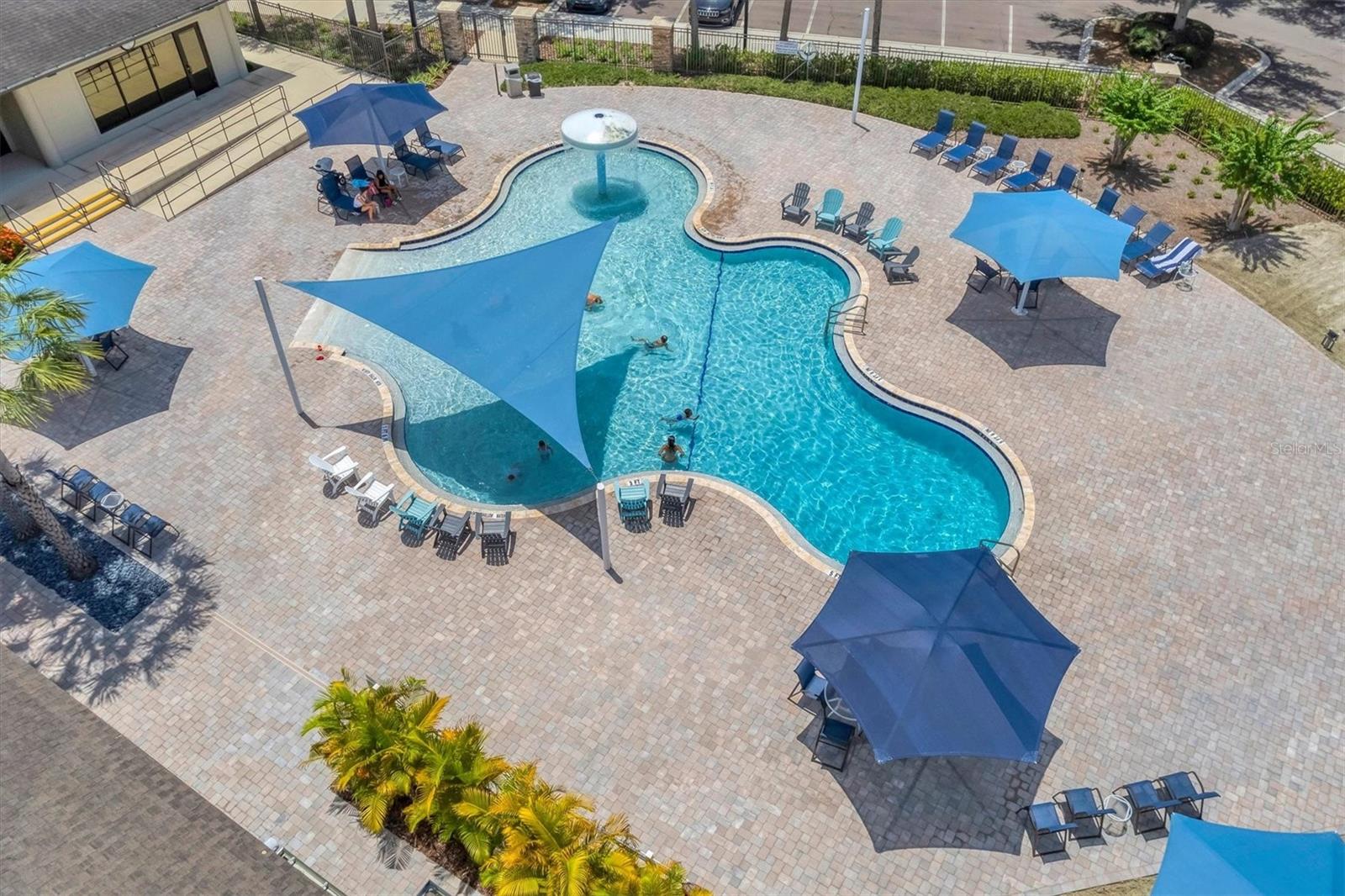 Aerial view of a curving swimming pool with a fountain, blue shade sails, and poolside loungers.