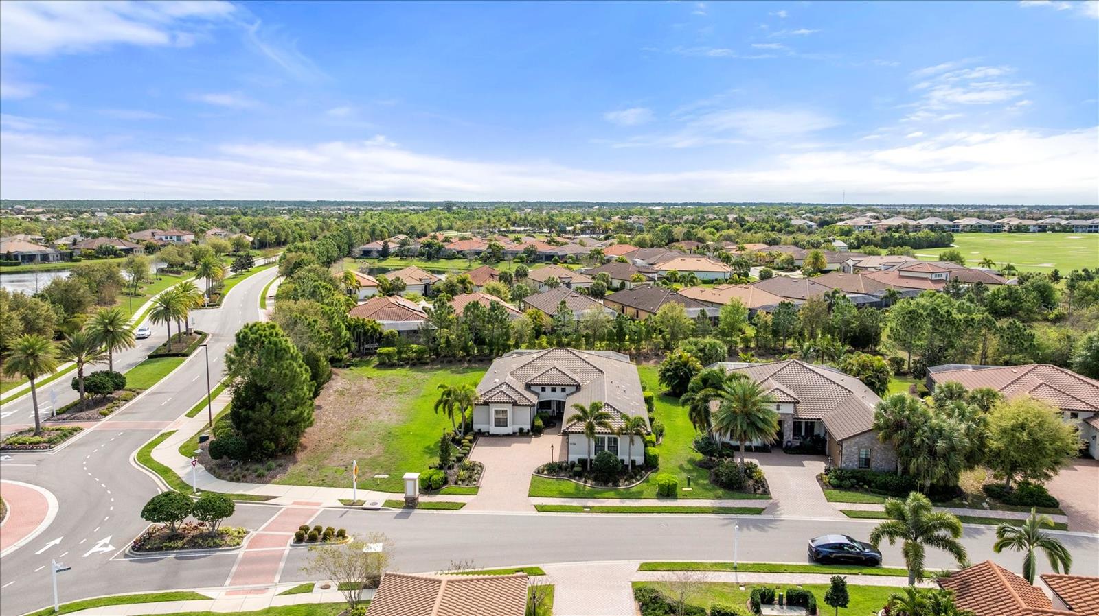 Aerial view of a tiled-roof home with landscaped grounds beside a curved, palm-lined neighborhood road.