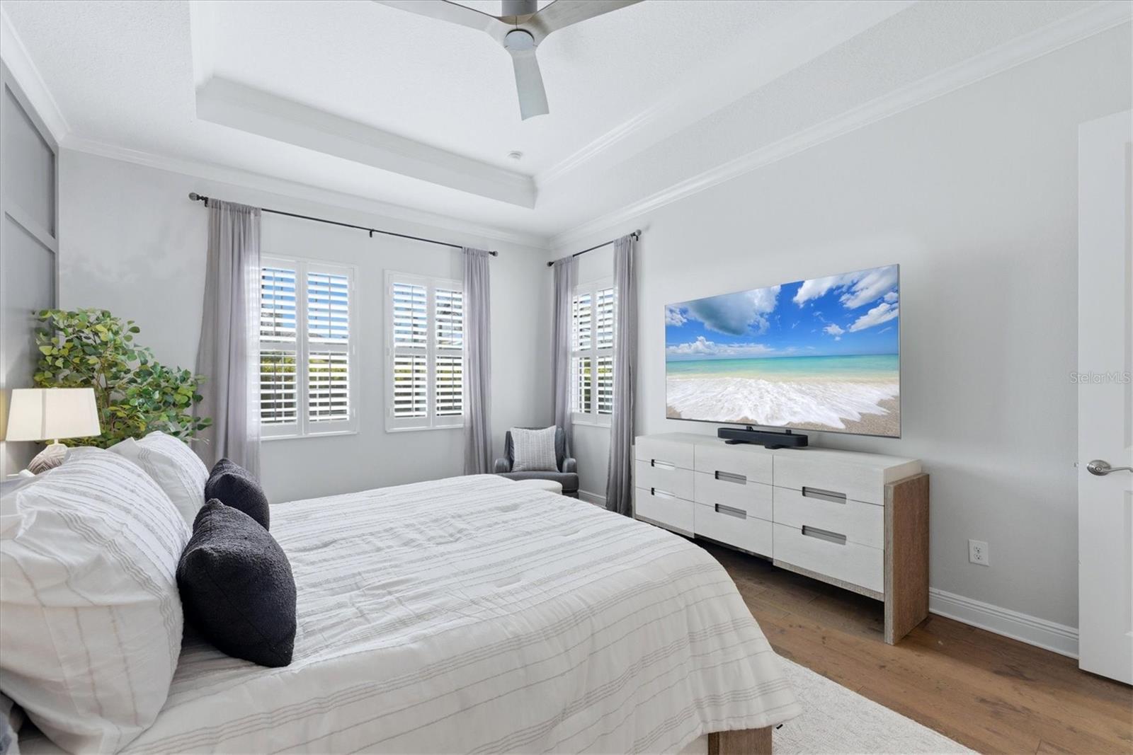 Bright bedroom with a white bed, tray ceiling, shuttered windows, and a dresser with a television.