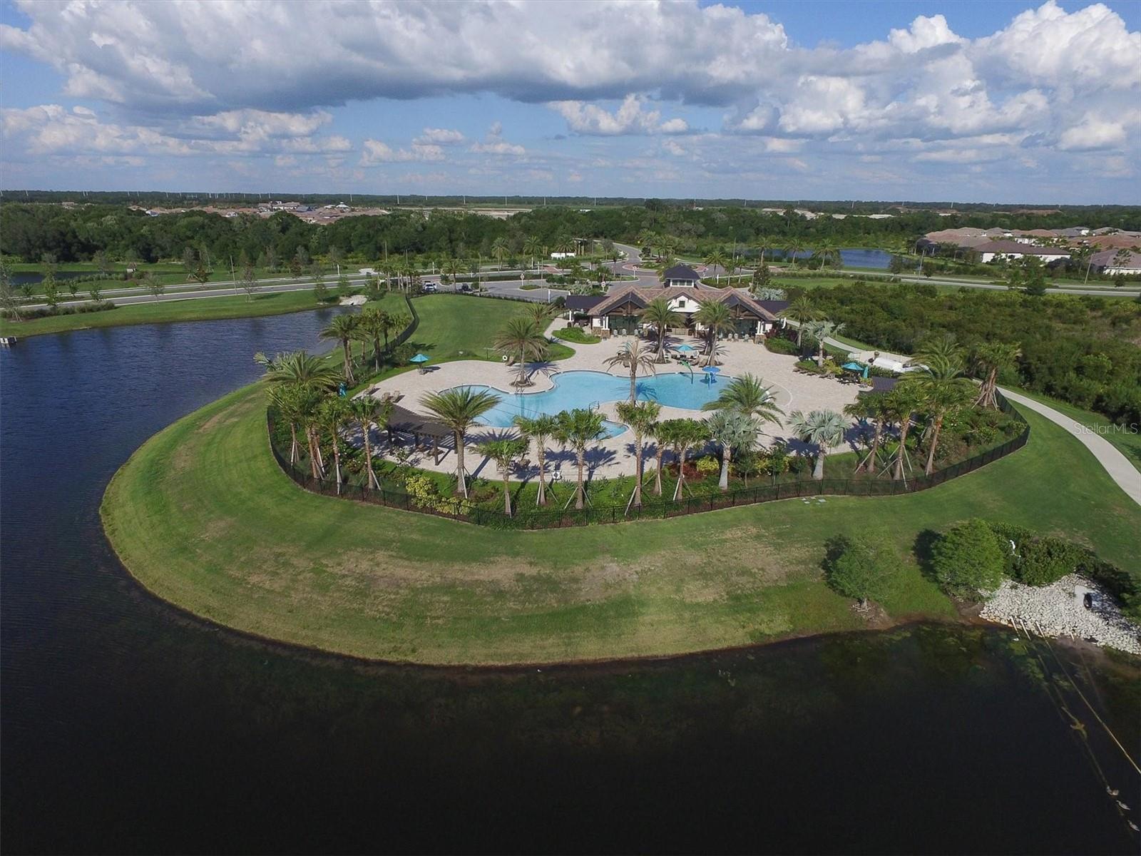 Aerial view of a community pool and clubhouse beside a pond, with palm trees and landscaped grounds.