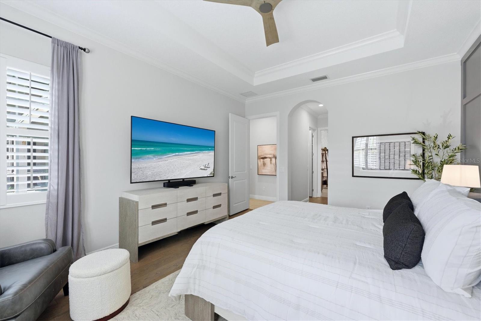 Bedroom with a white bed, tray ceiling, plantation shutters, and a wall-mounted television showing a beach.