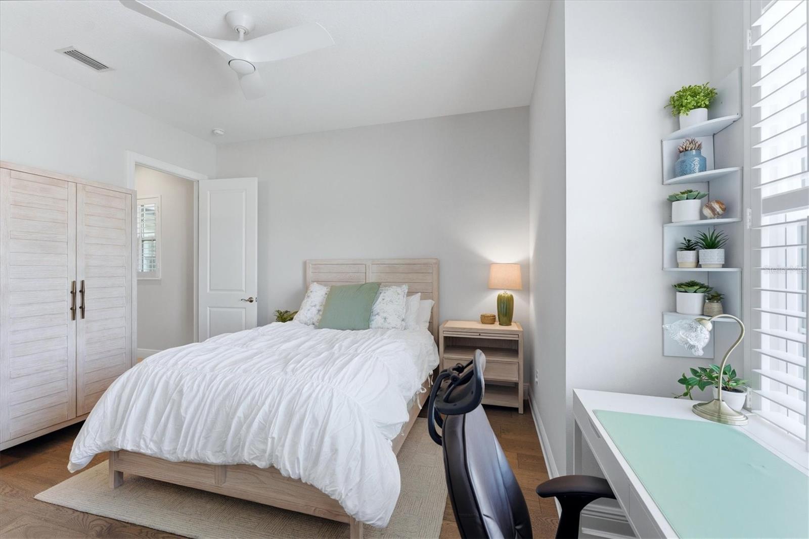 Furnished bedroom with a light wood bed, louvered wardrobe, and desk beside shuttered windows.
