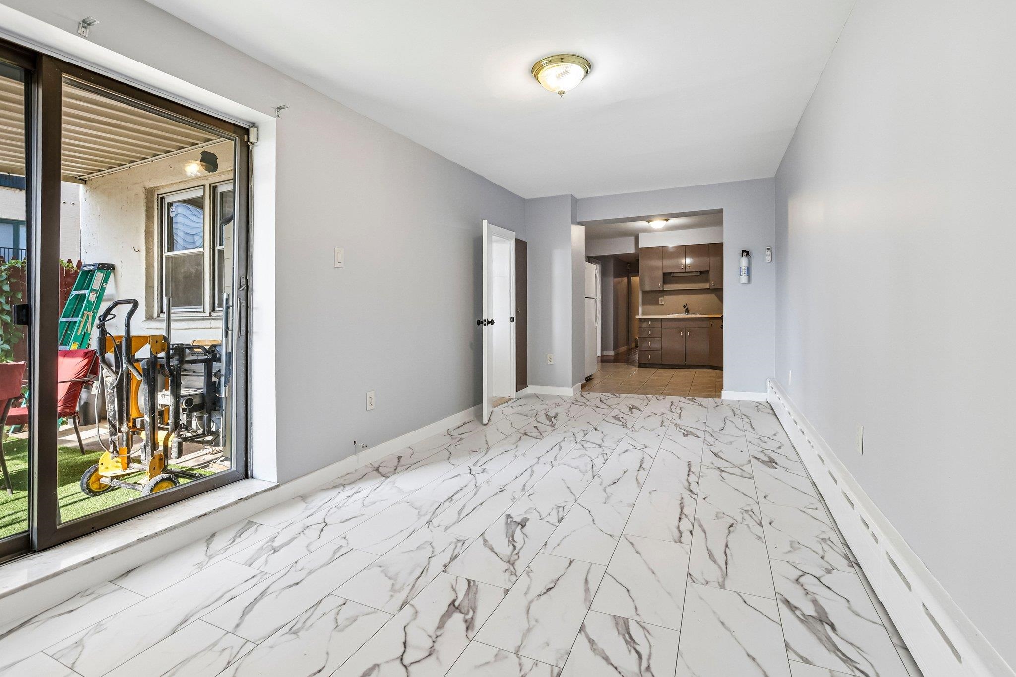 Empty living area with marble-look tile floors, sliding glass patio doors, and a kitchen beyond.