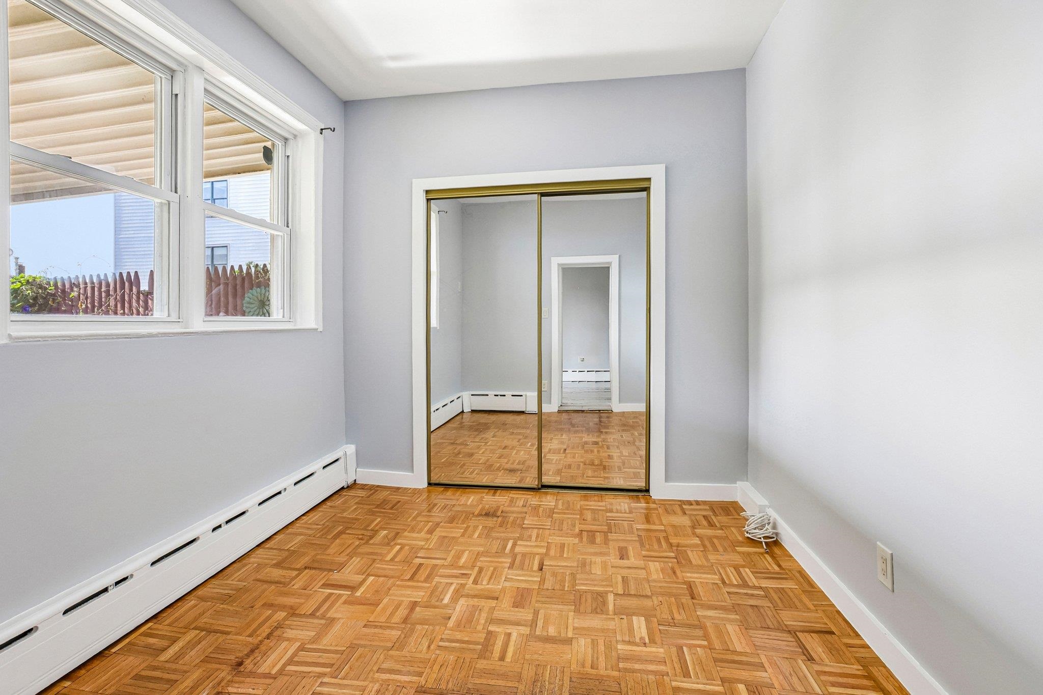 Vacant room with parquet floors, large windows, baseboard heating, and mirrored sliding closet doors.