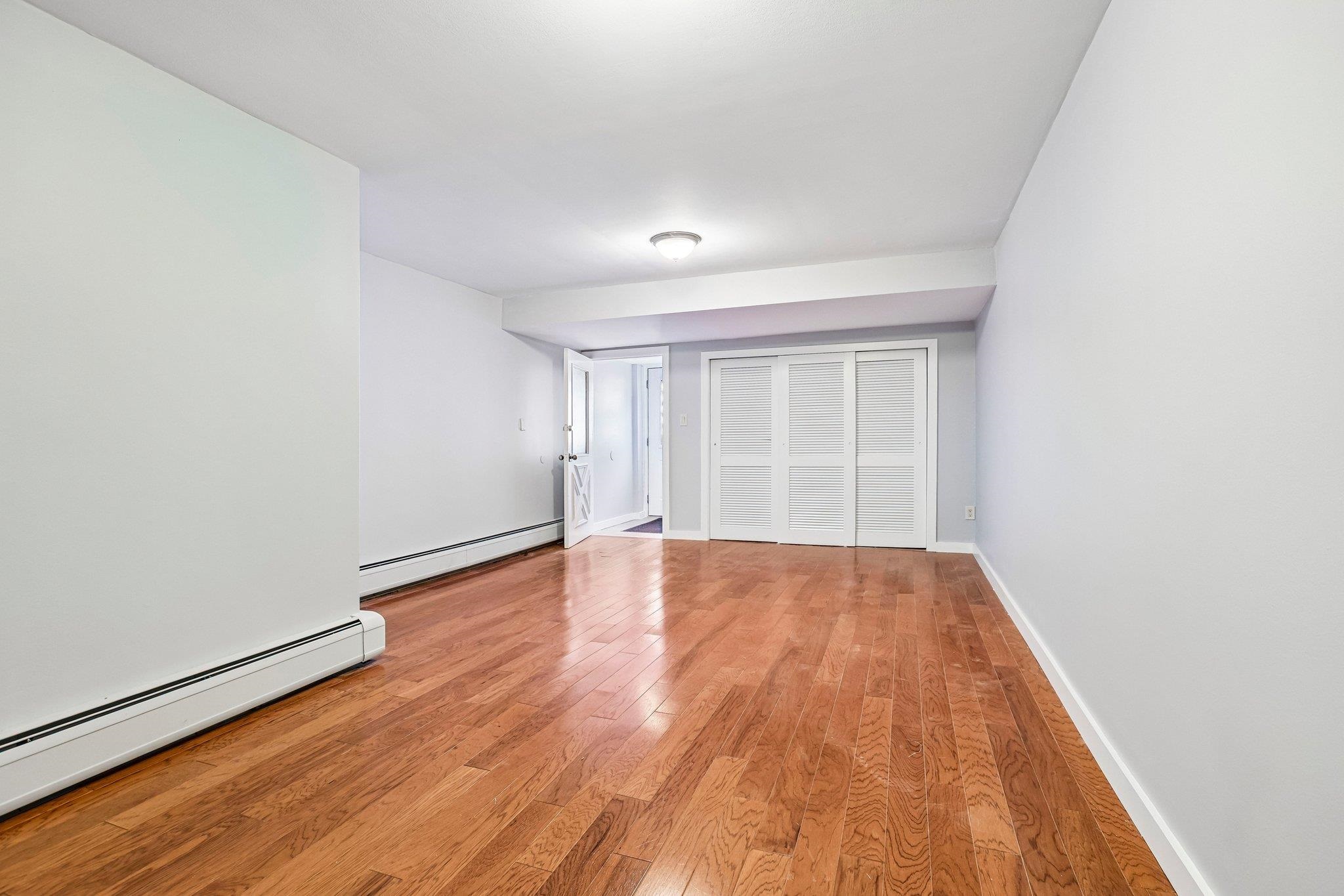 Empty room with reddish hardwood floors, baseboard heating, and white louvered closet doors.