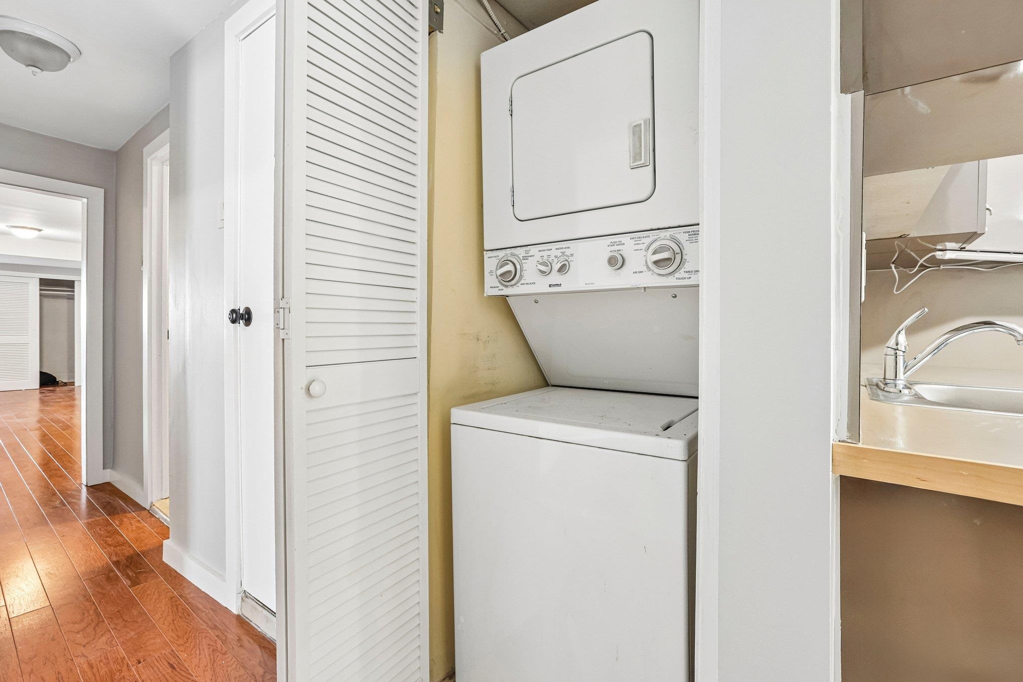 Stacked washer and dryer in a laundry nook beside a kitchen sink and wood-floored hallway.