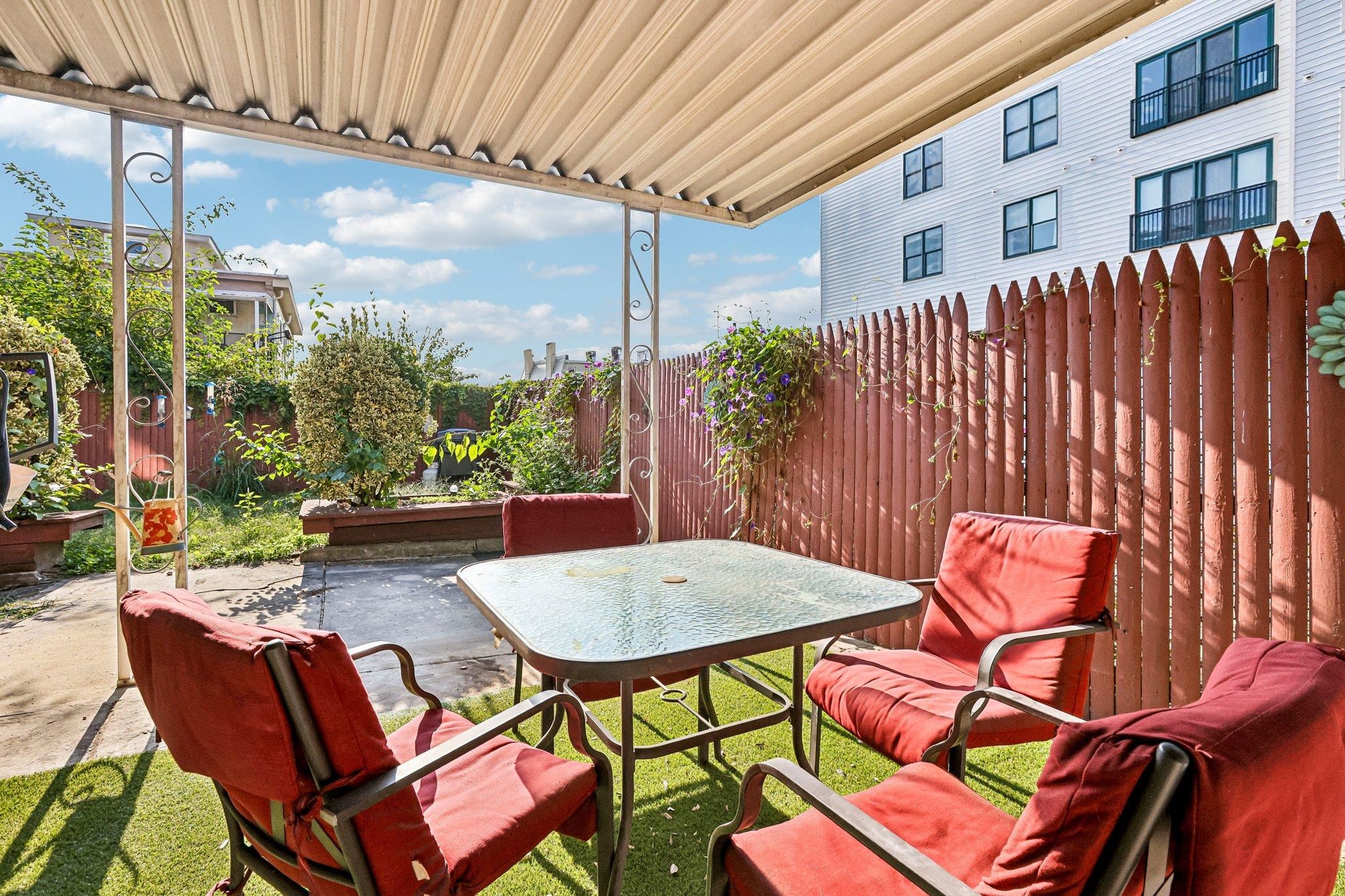 Covered patio with a glass-top table, red-cushioned chairs, garden beds, and a tall wooden fence.