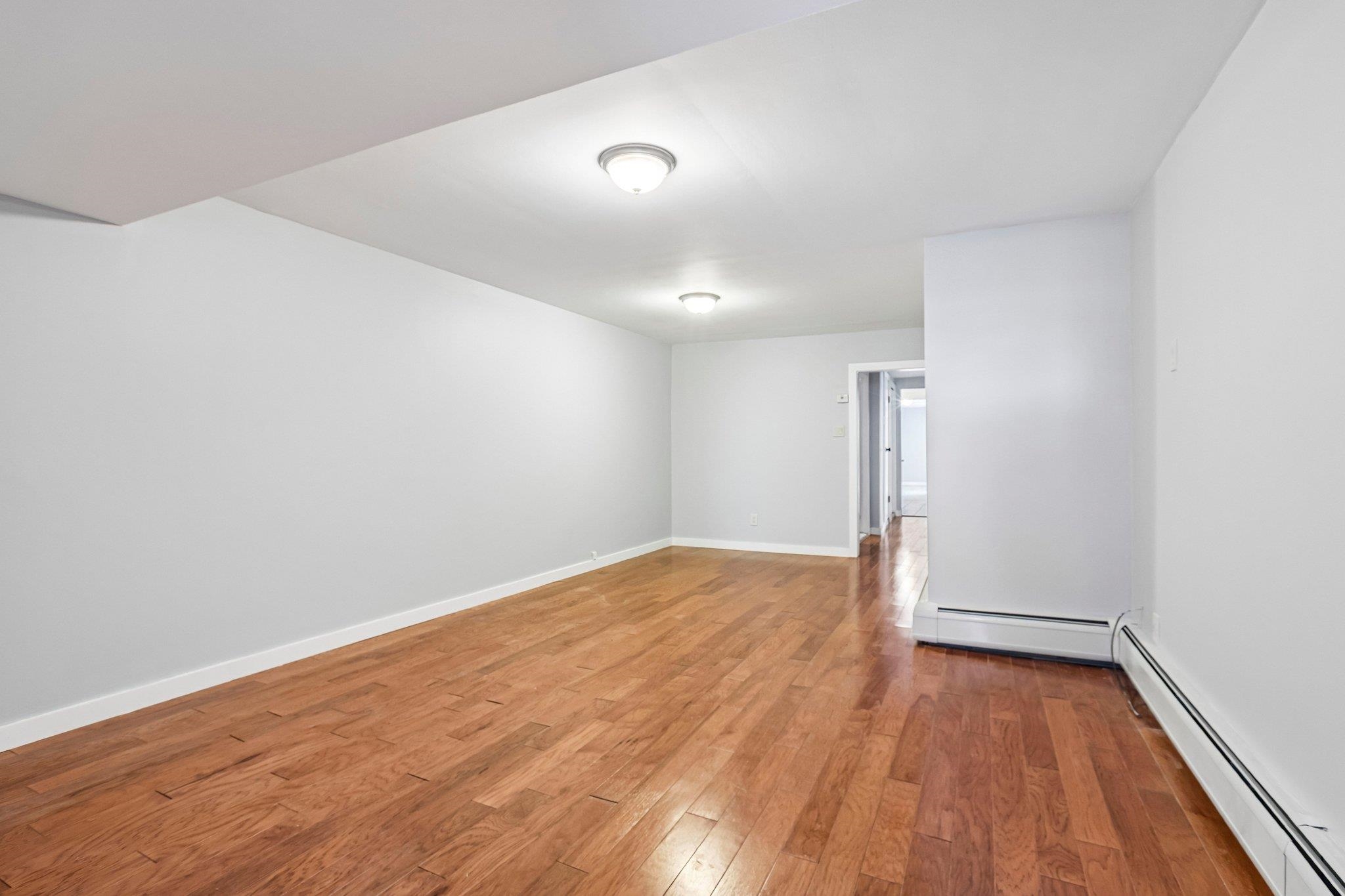An empty living room with hardwood floors, white walls, ceiling lights, and a hallway doorway.