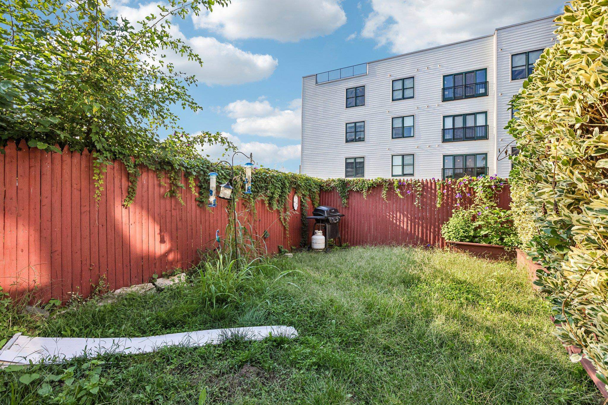 Grassy backyard with a red fence, leafy vines, planters, bird feeders, and a barbecue grill.