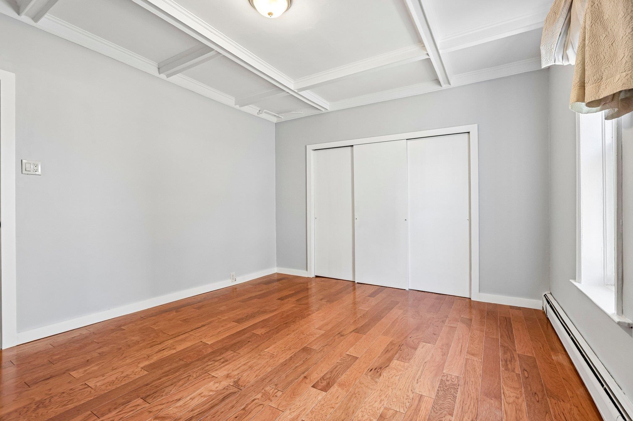 Empty bedroom with hardwood floors, a coffered ceiling, sliding closet doors, and a curtained window.