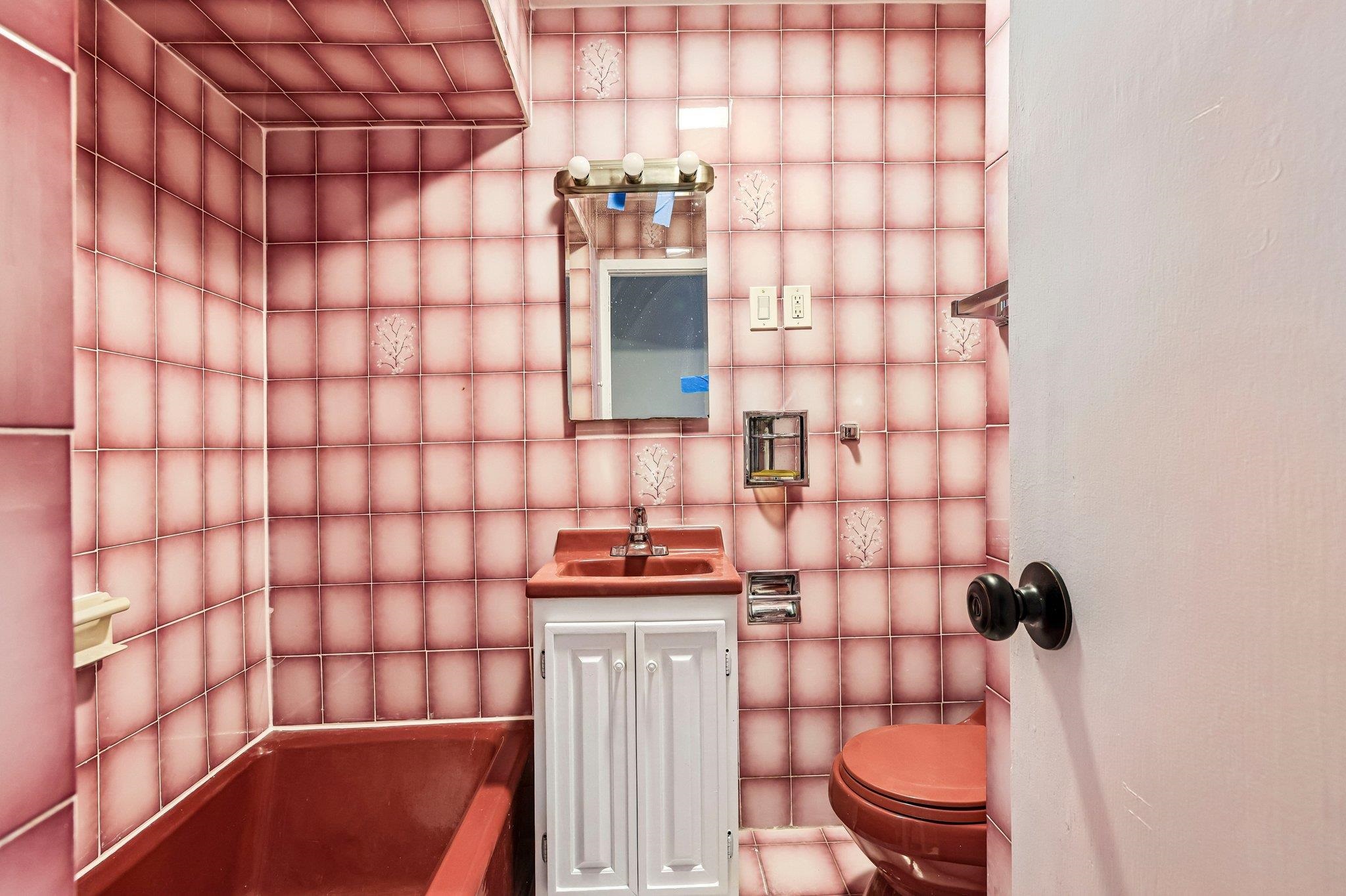 Bathroom with pink patterned tile, reddish bathtub and toilet, and white vanity.