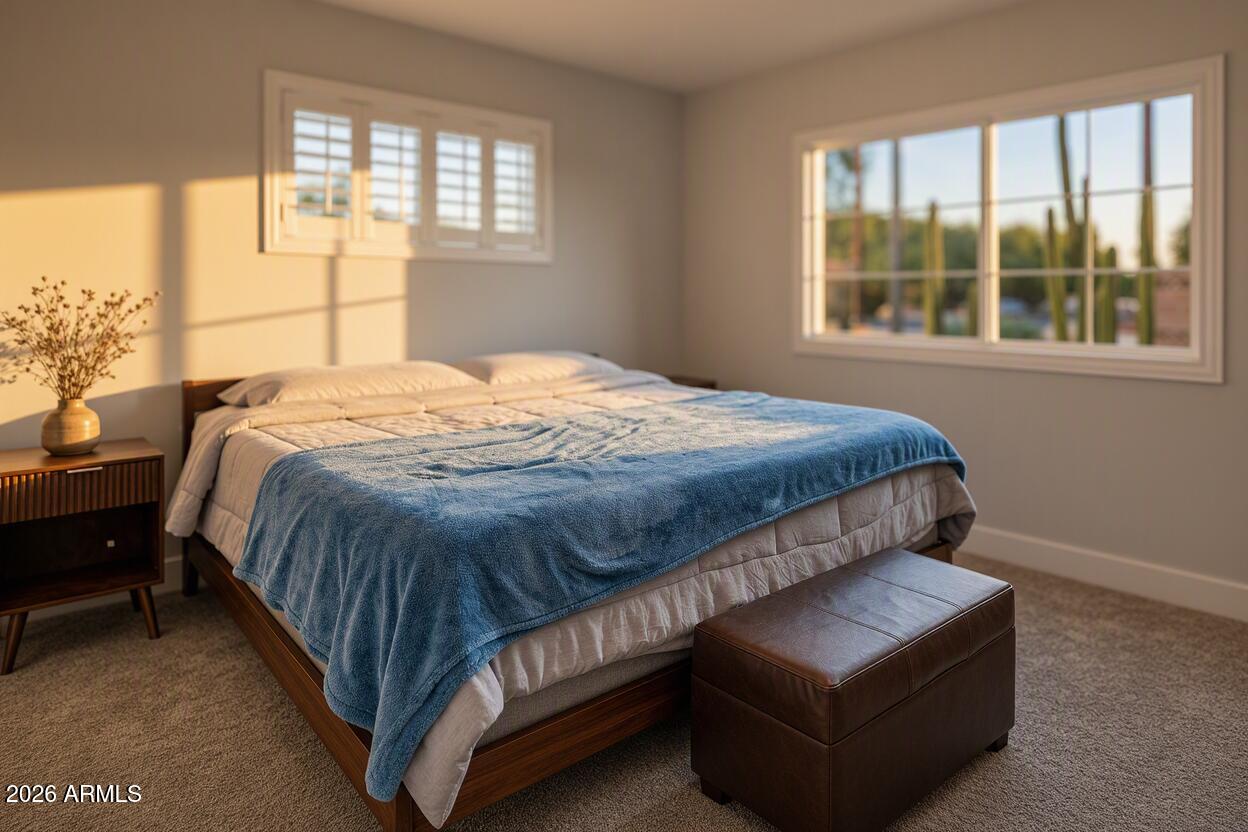 Furnished bedroom with a blue blanket, leather ottoman, and large window overlooking desert landscaping.