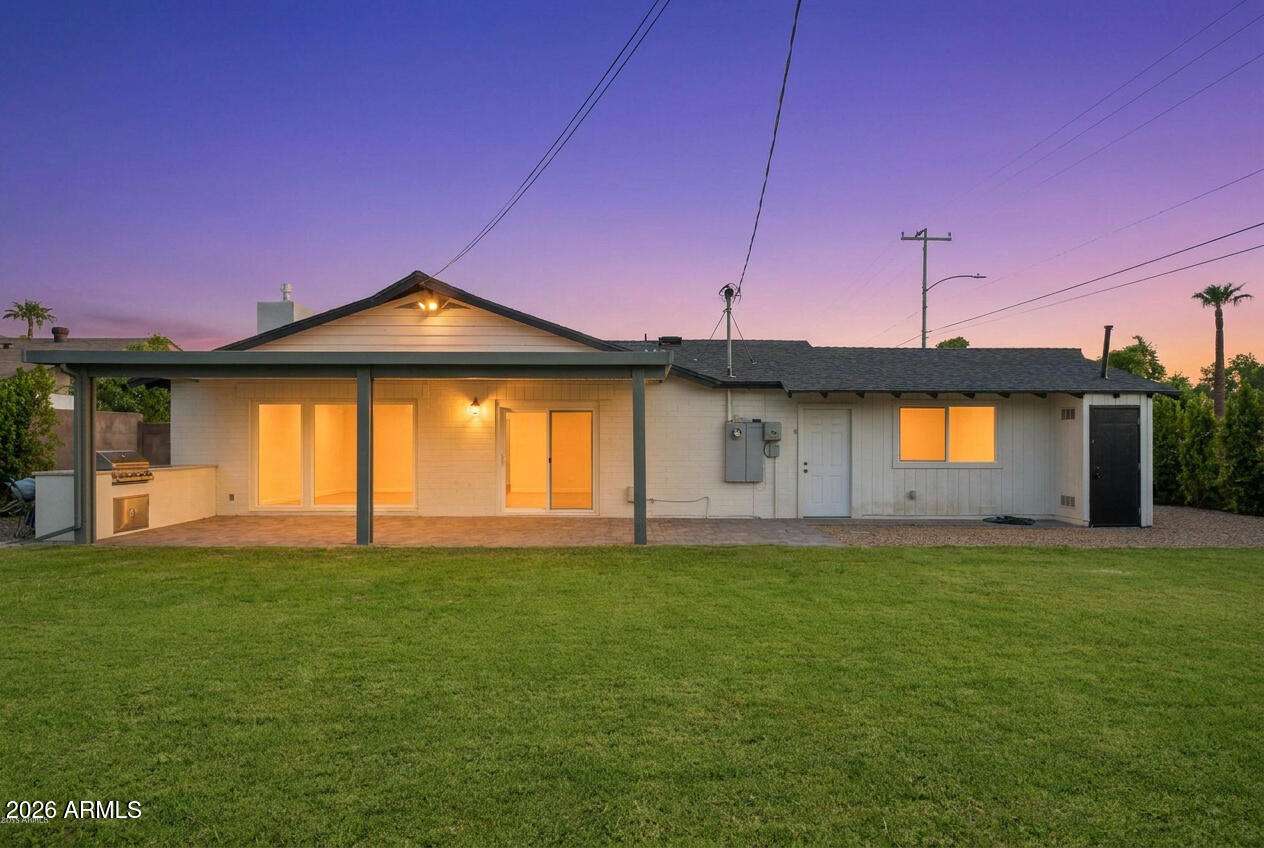 Rear of a single-story home with a covered patio, outdoor grill, and lawn at dusk.
