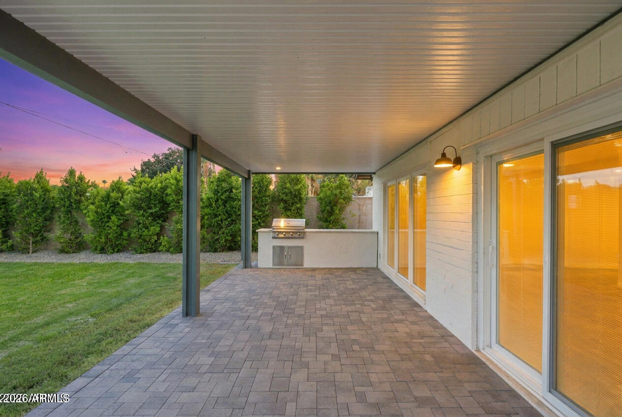 Covered backyard patio with paver flooring, built-in grill, and sliding glass doors at dusk.