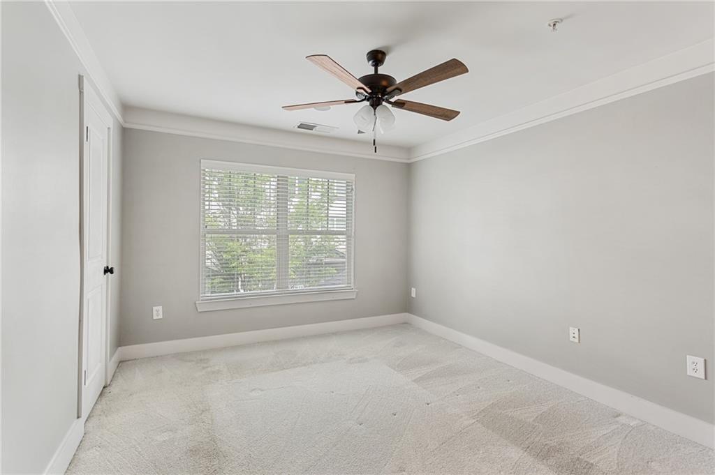 Empty bedroom with a wide blinded window, pale carpet, and a wood-blade ceiling fan.