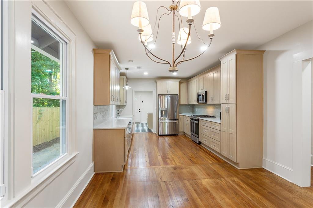 Kitchen with light wood cabinetry, stainless steel appliances, hardwood floors, and a large window overlooking a fenced yard.
