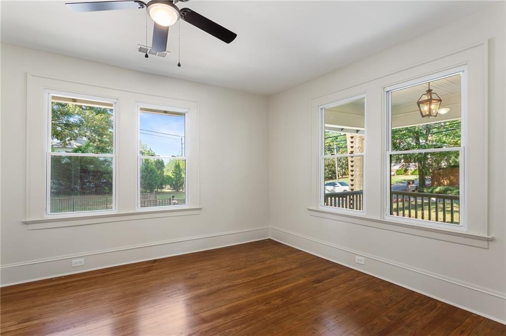Empty room with hardwood floors, ceiling fan, and windows overlooking trees and a porch.