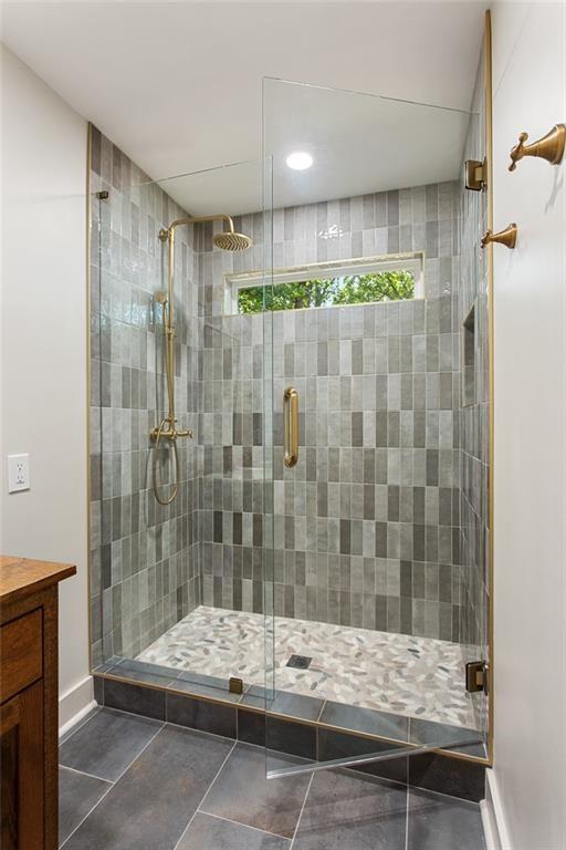 Walk-in shower with gray tile, brass fixtures, a pebble-patterned floor, and a narrow window.