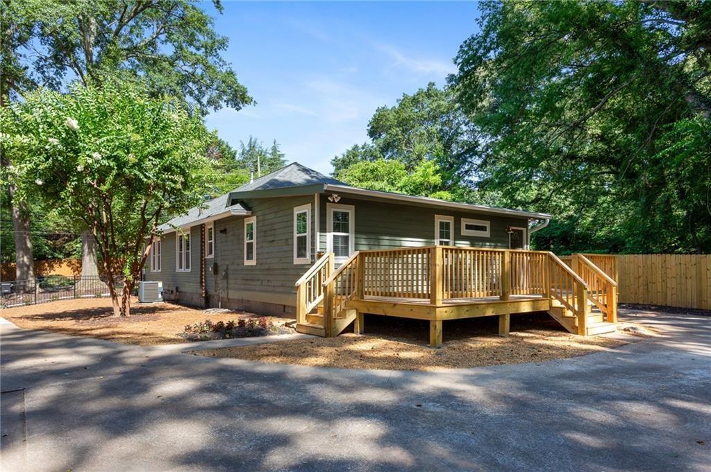 Gray single-story house with a raised wooden deck, mature trees, and a broad paved driveway.