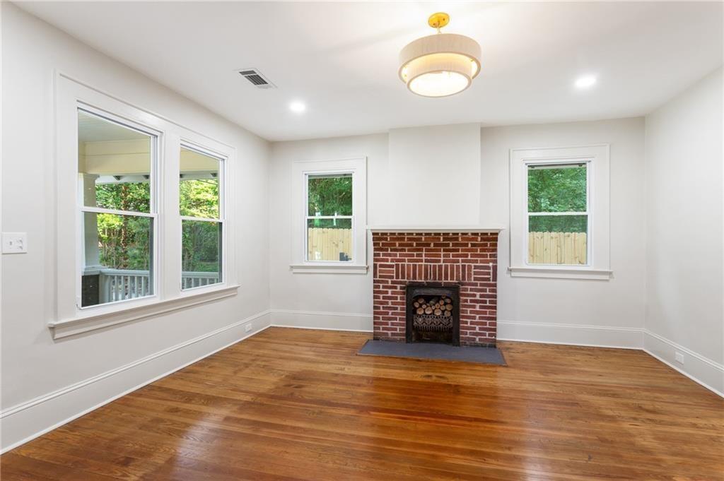 Vacant living room with hardwood floors, a red brick fireplace, and windows overlooking greenery.