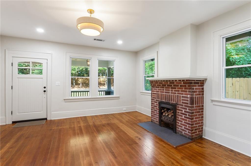 Empty living room with hardwood floors, a brick fireplace, and windows overlooking greenery.