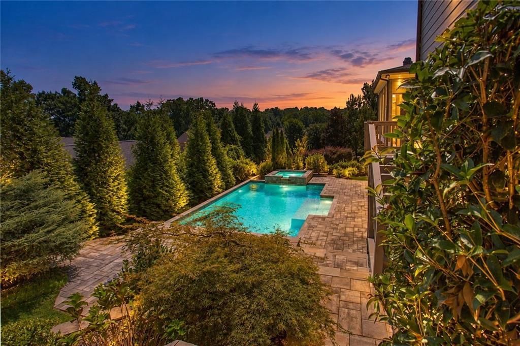 Backyard pool and raised spa beside a stone patio, framed by dense landscaping at sunset.