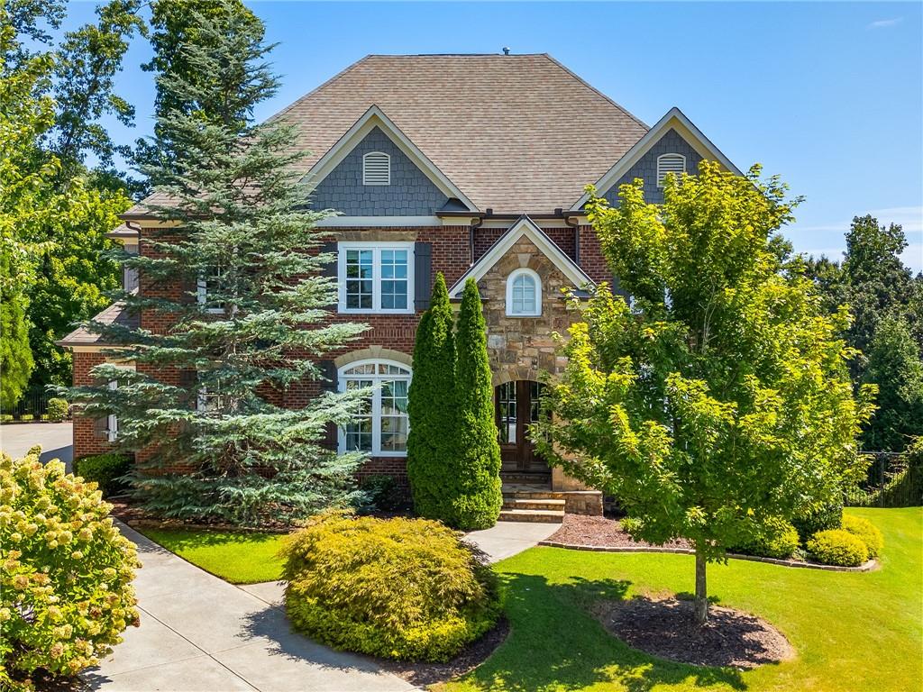 Two-story brick and stone home framed by mature trees, with a landscaped lawn and concrete driveway.