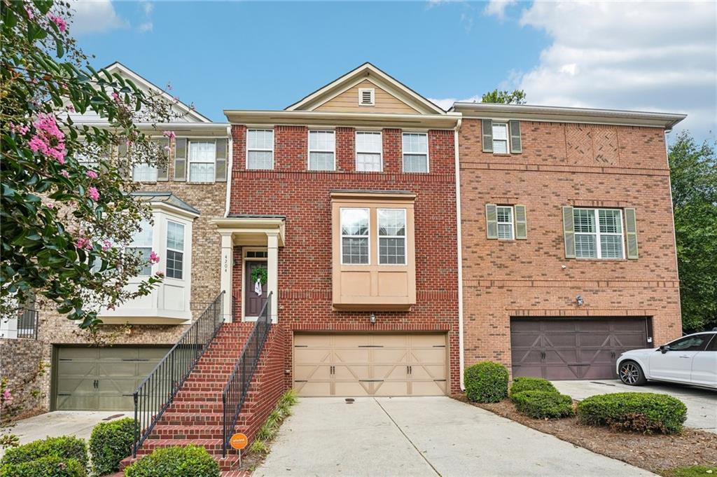 Attached brick townhomes with front stairways, garage doors, and flowering landscaping.