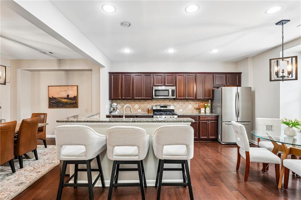 Open kitchen with a granite island, three upholstered stools, dark cabinets, and stainless steel appliances.