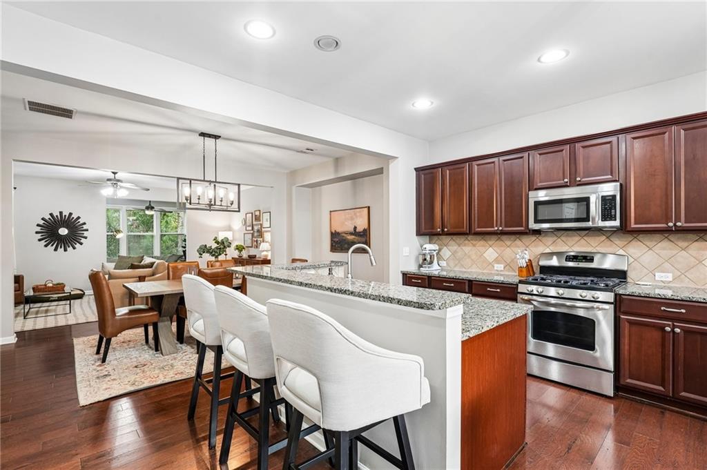 Open-plan kitchen with granite island, white bar stools, dark wood cabinets, and stainless steel appliances.