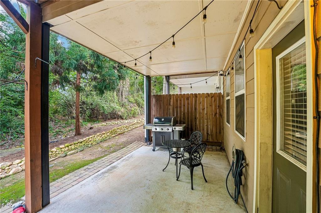 Covered patio with string lights, grill, and metal seating beside wooded greenery.