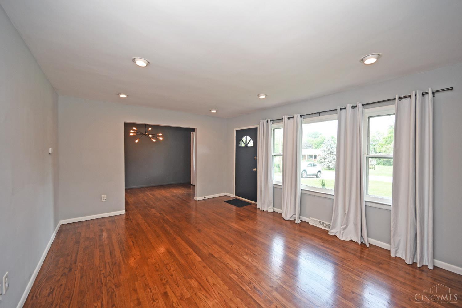 Vacant living room with polished hardwood floors, curtained windows, and an open doorway to a dining room.