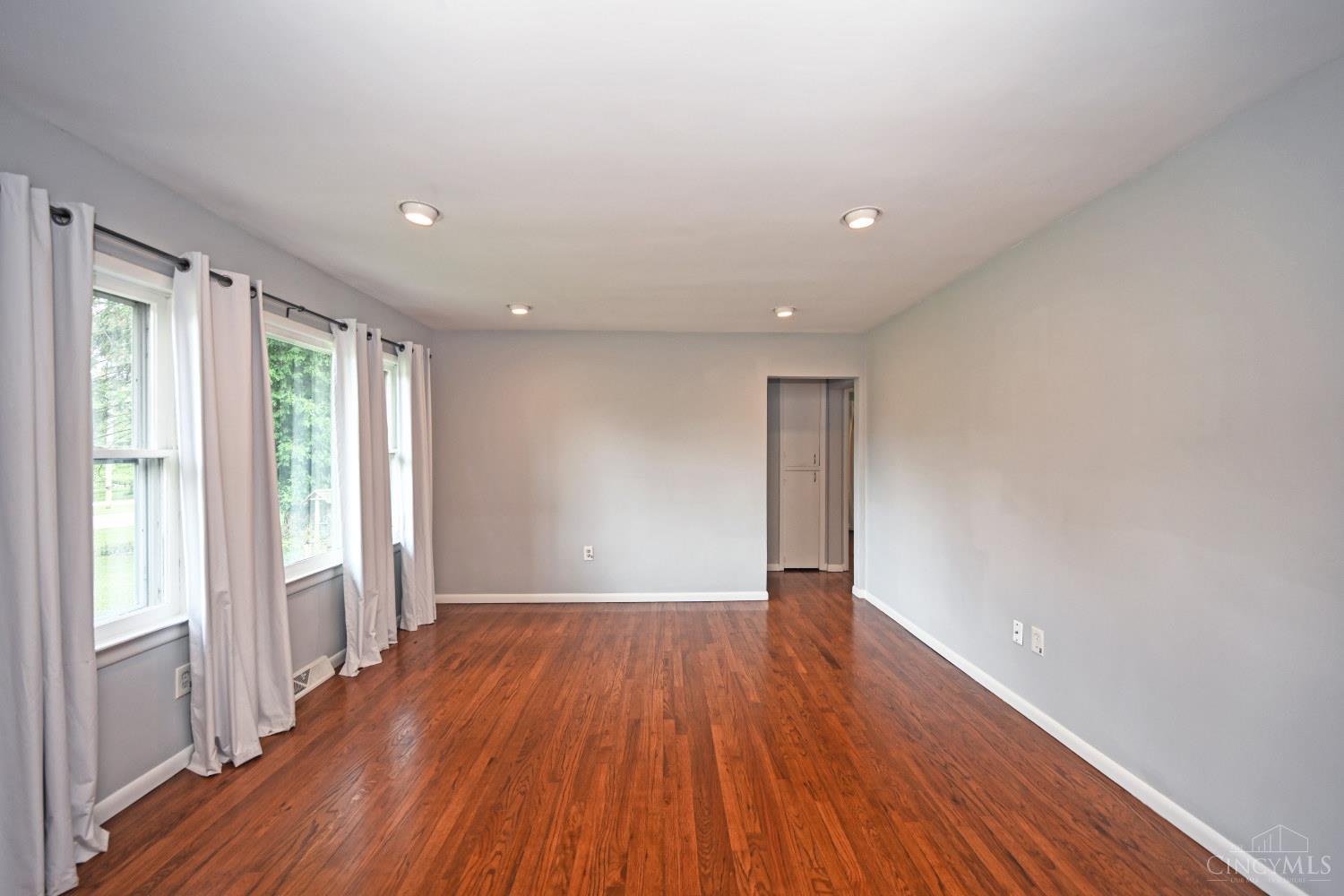 Empty living room with hardwood floors and curtained windows along one wall.