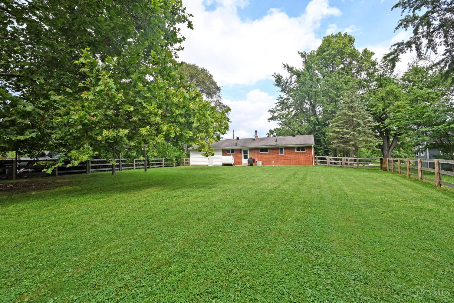 Rear of a brick-and-siding ranch home beyond a broad fenced lawn and mature trees.