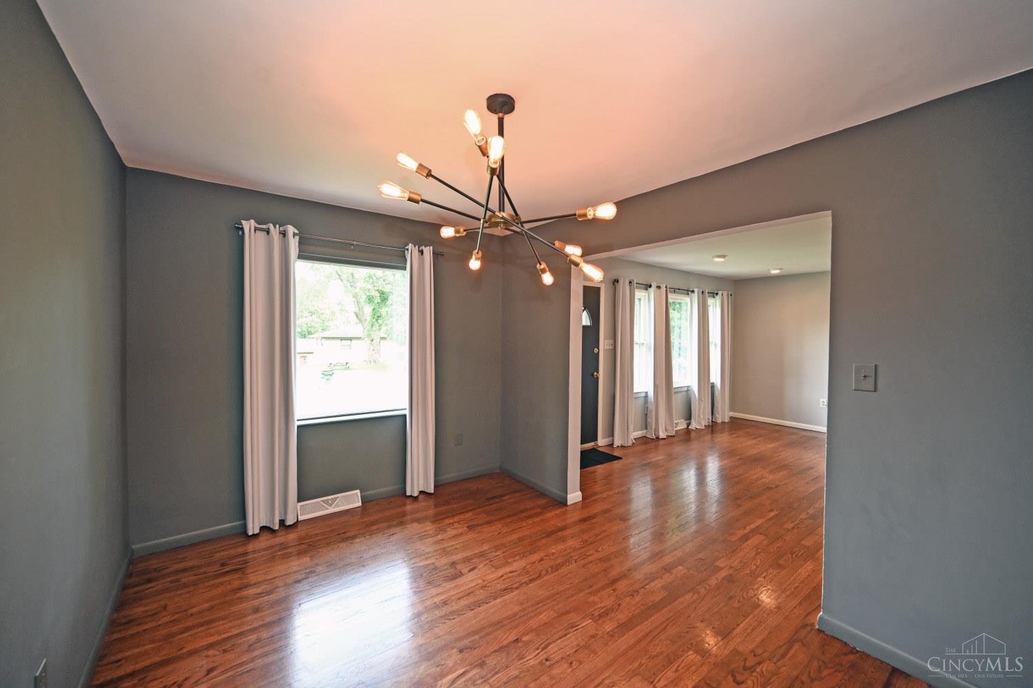 Empty dining area with hardwood floors, gray walls, and a modern chandelier opening into a bright adjoining room.