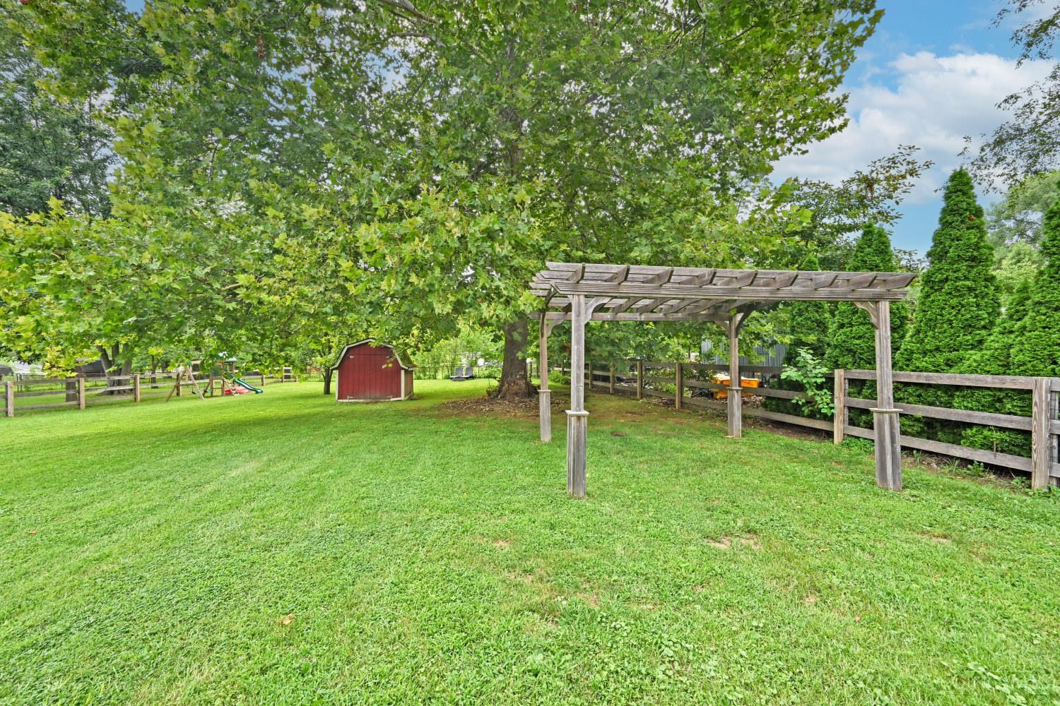 Tree-shaded backyard with a wooden pergola, red shed, playset, and broad lawn.