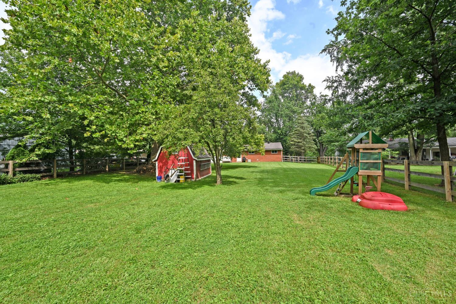 Fenced backyard with a red shed, mature trees, and a wooden playset with a green slide.