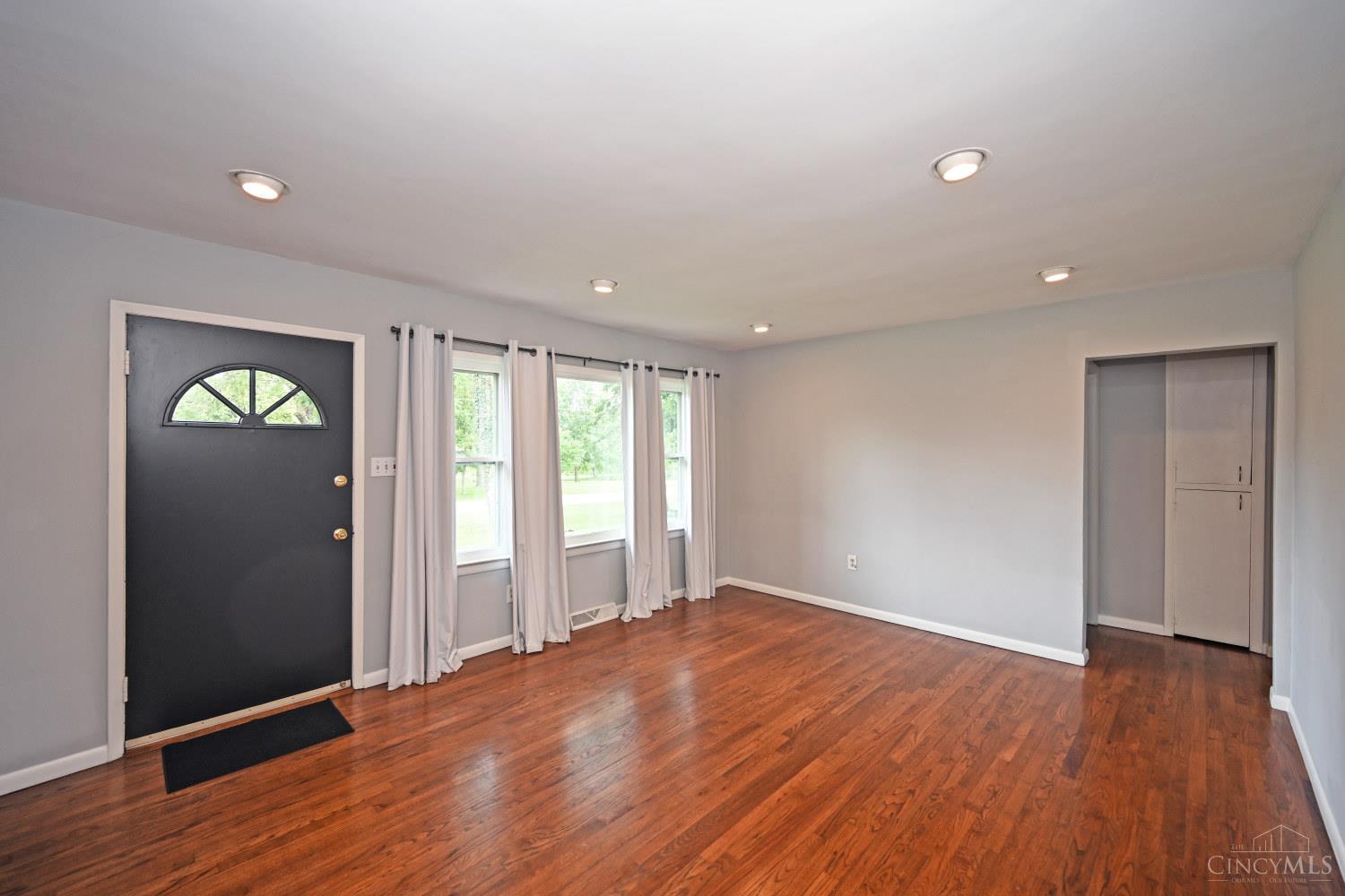 Vacant living room with reddish hardwood floors, curtained windows, and a black entry door.