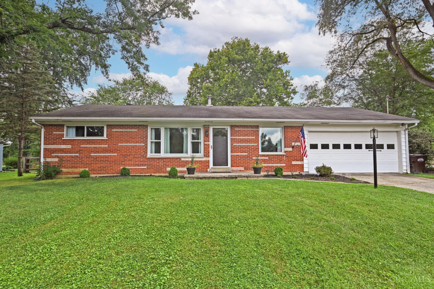 Red brick ranch home with an attached white garage, a green lawn, and mature trees.
