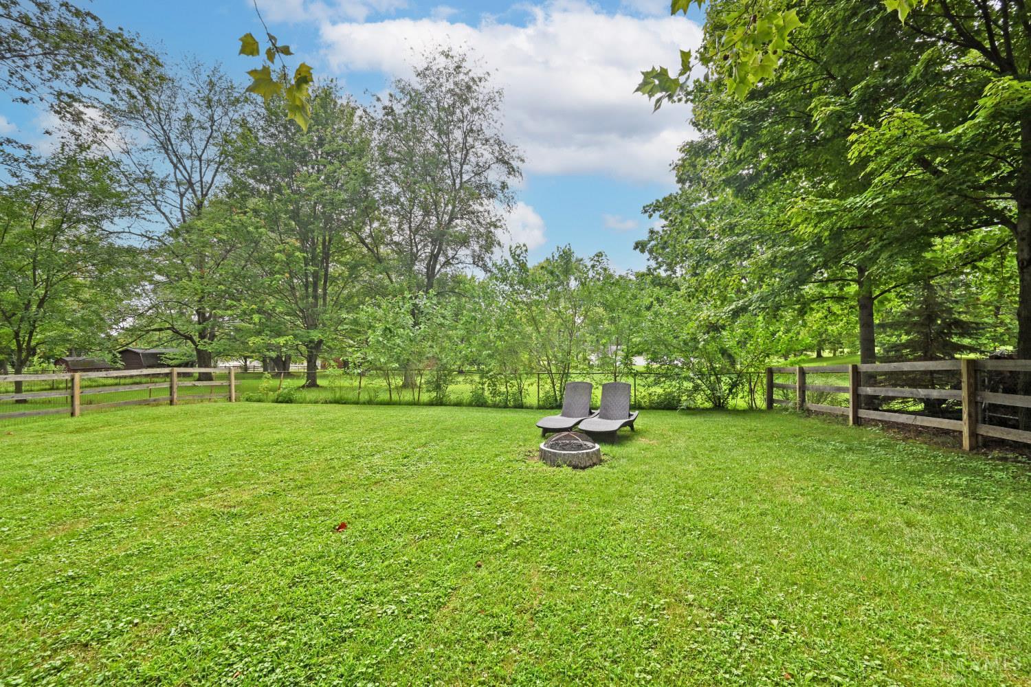 Fenced backyard with a wide green lawn, mature trees, lounge chairs, and a stone fire pit.