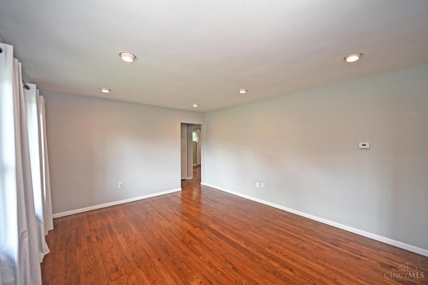 Vacant living room with hardwood floors, pale walls, and curtained windows.