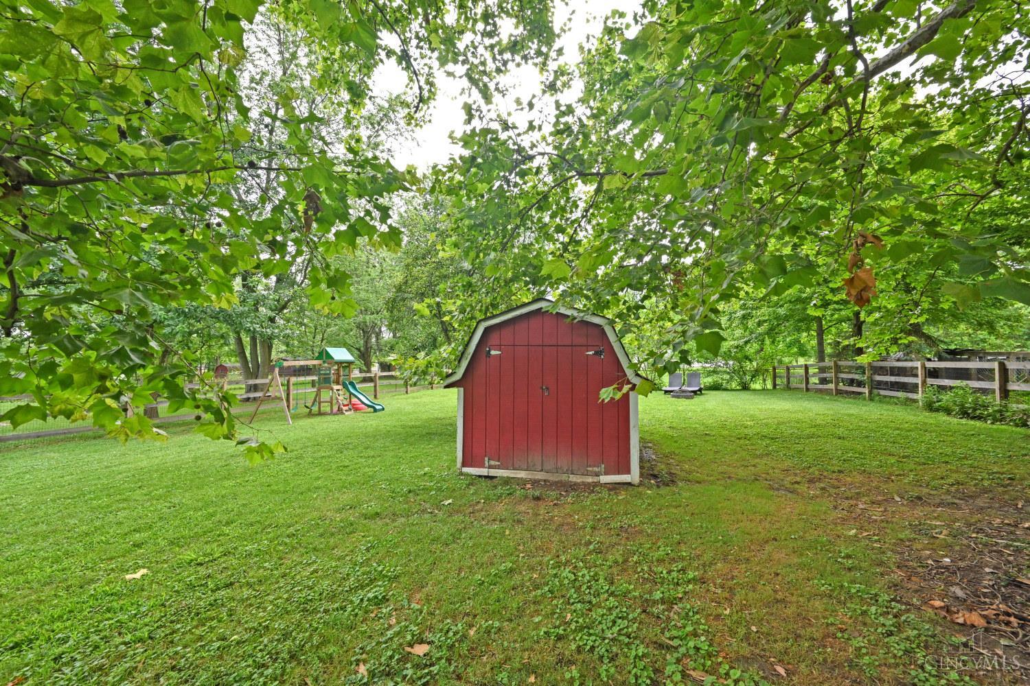 Backyard with a red storage shed, fenced lawn, and children’s playset beneath mature trees.